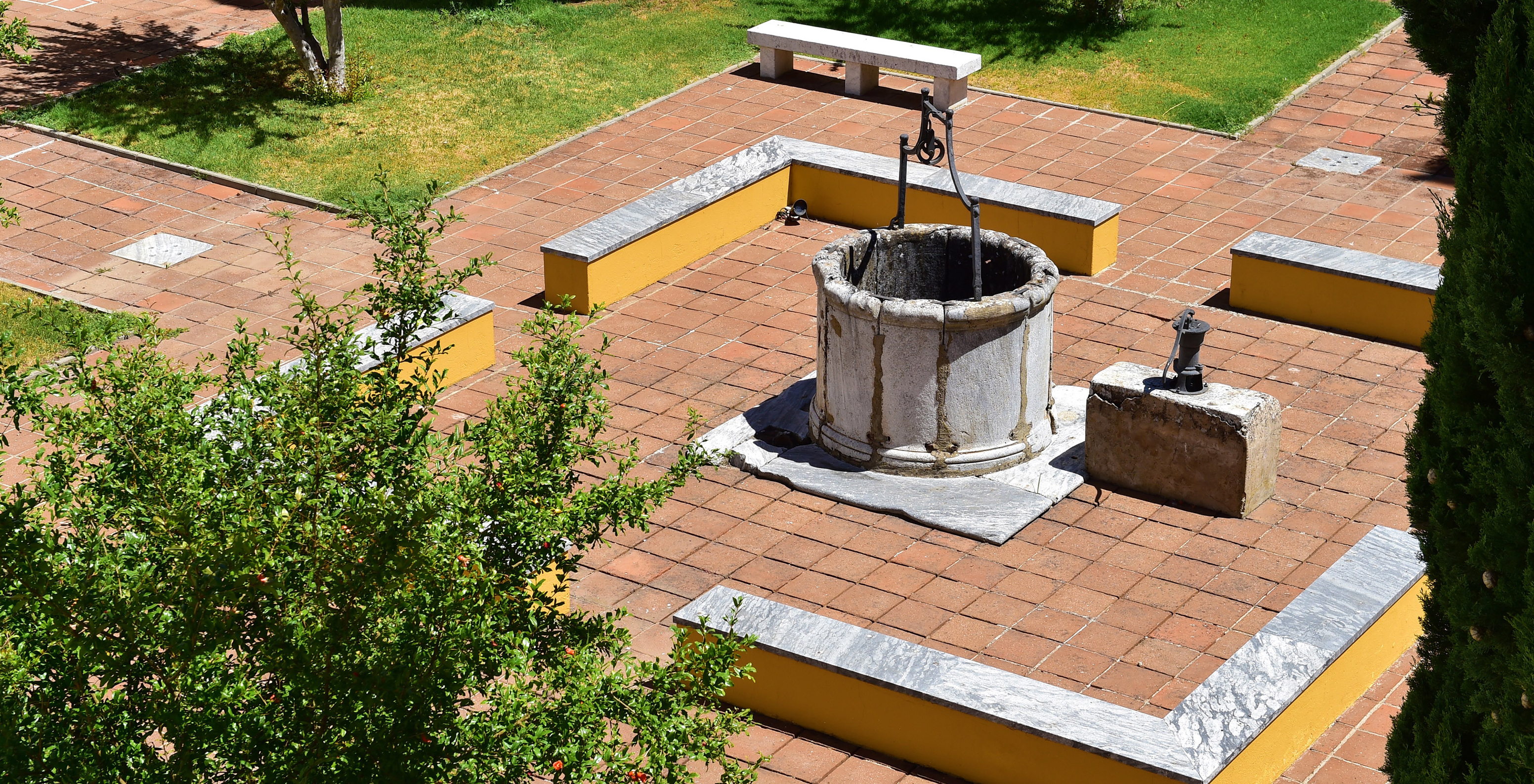 Brunnen in der Pousada Convento Beja mit Bänken darum herum und einem Garten mit Bäumen und Gras, das das Bild ergänzt