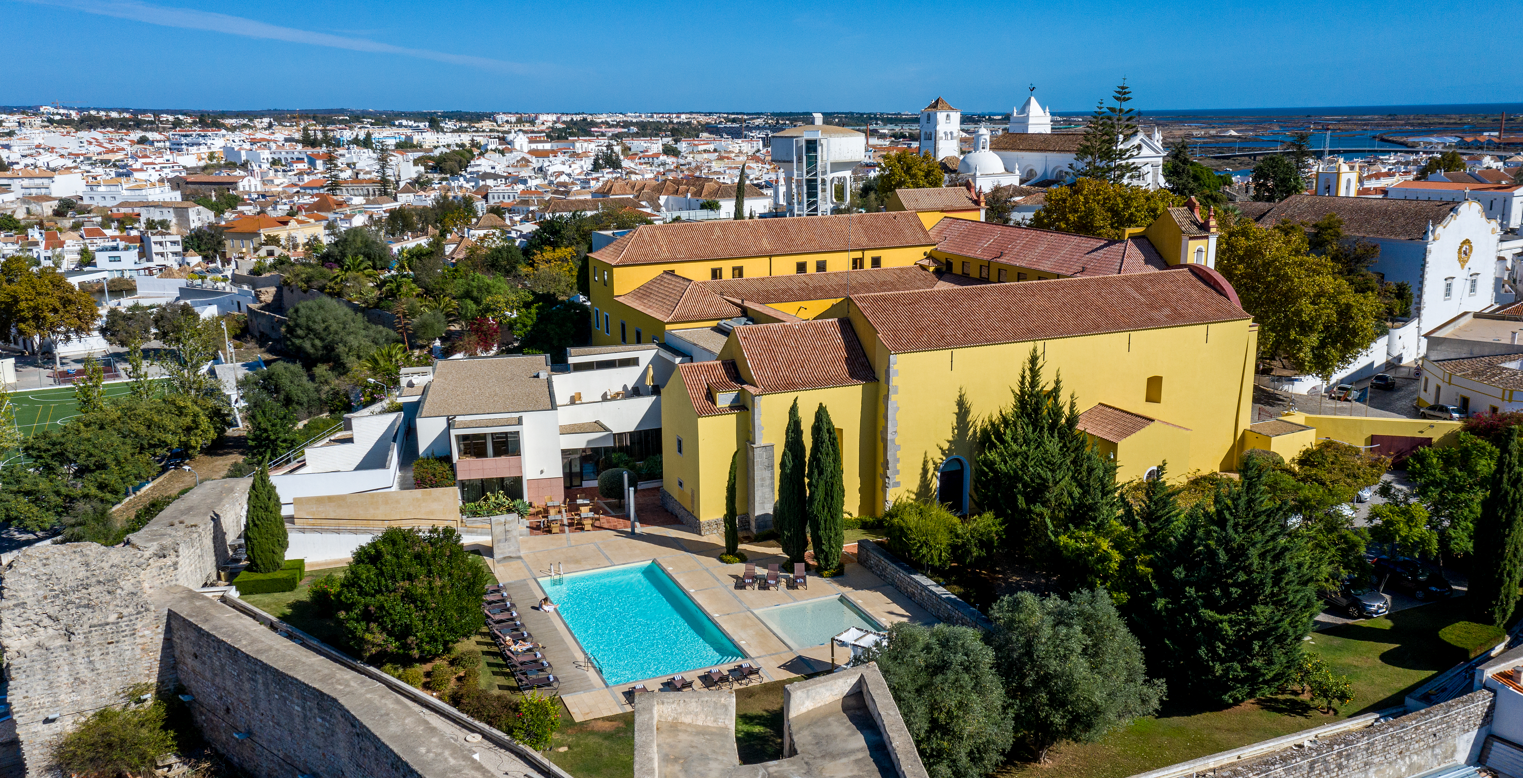 Luftaufnahme der Pousada Convento de Tavira mit dem gelben Gebäude, dem Außenpool und der Stadt im Hintergrund