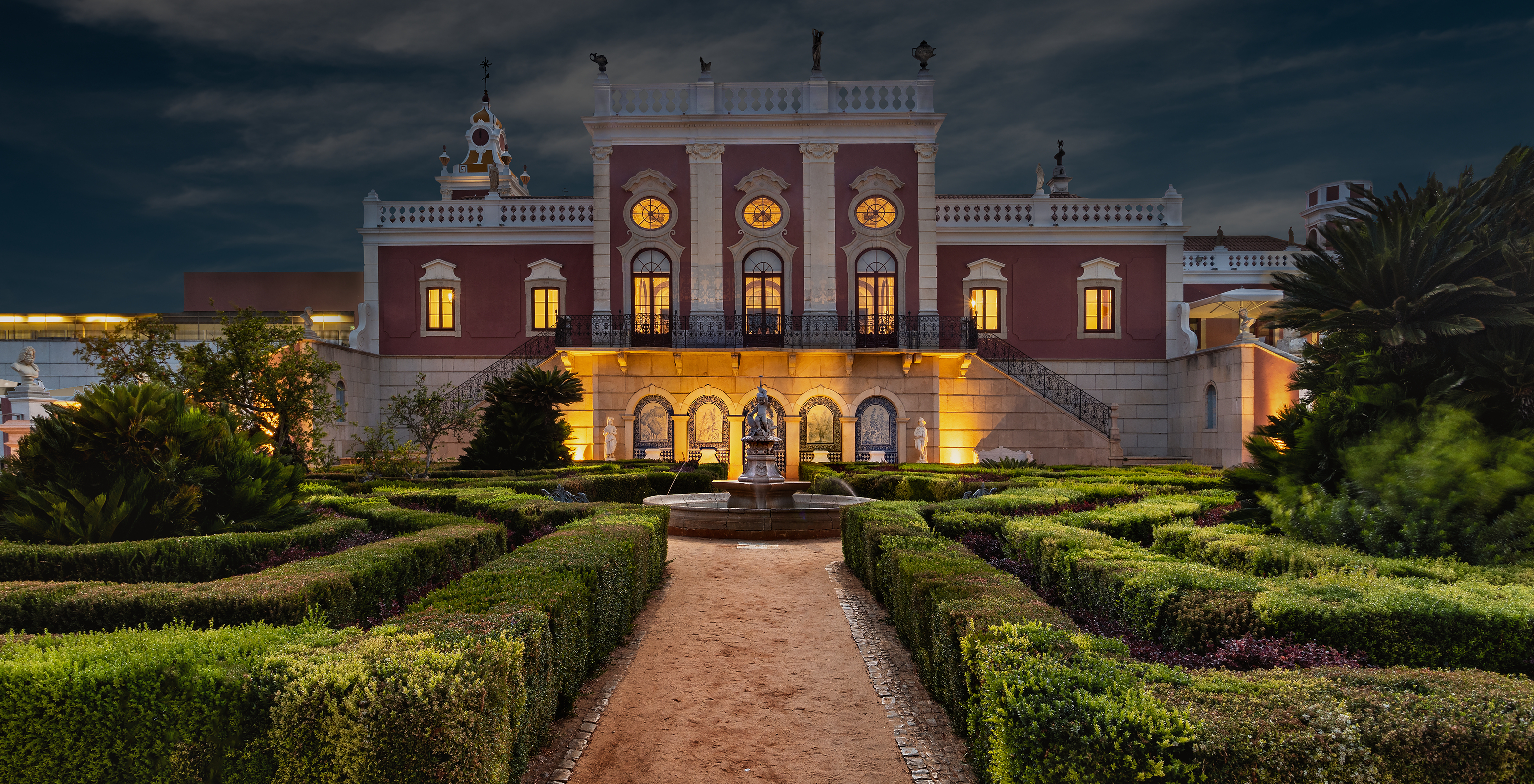 Fassade bei Nacht der Pousada Palácio Estoi, einem historischen Hotel in Faro, beleuchtet mit gepflegten Gärten und Brunnen