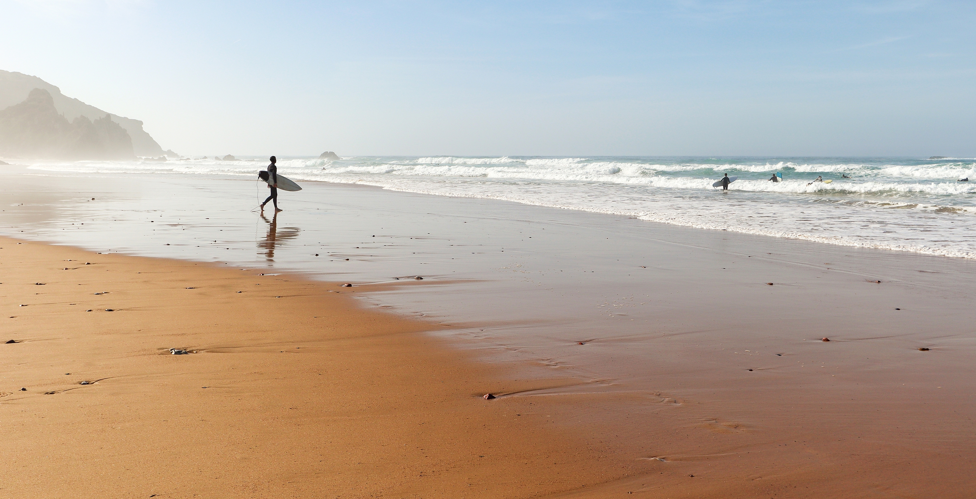 Einsamer Surfer am Wasser am Strand von Sagres mit einigen Wellen und anderen Surfern im Meer