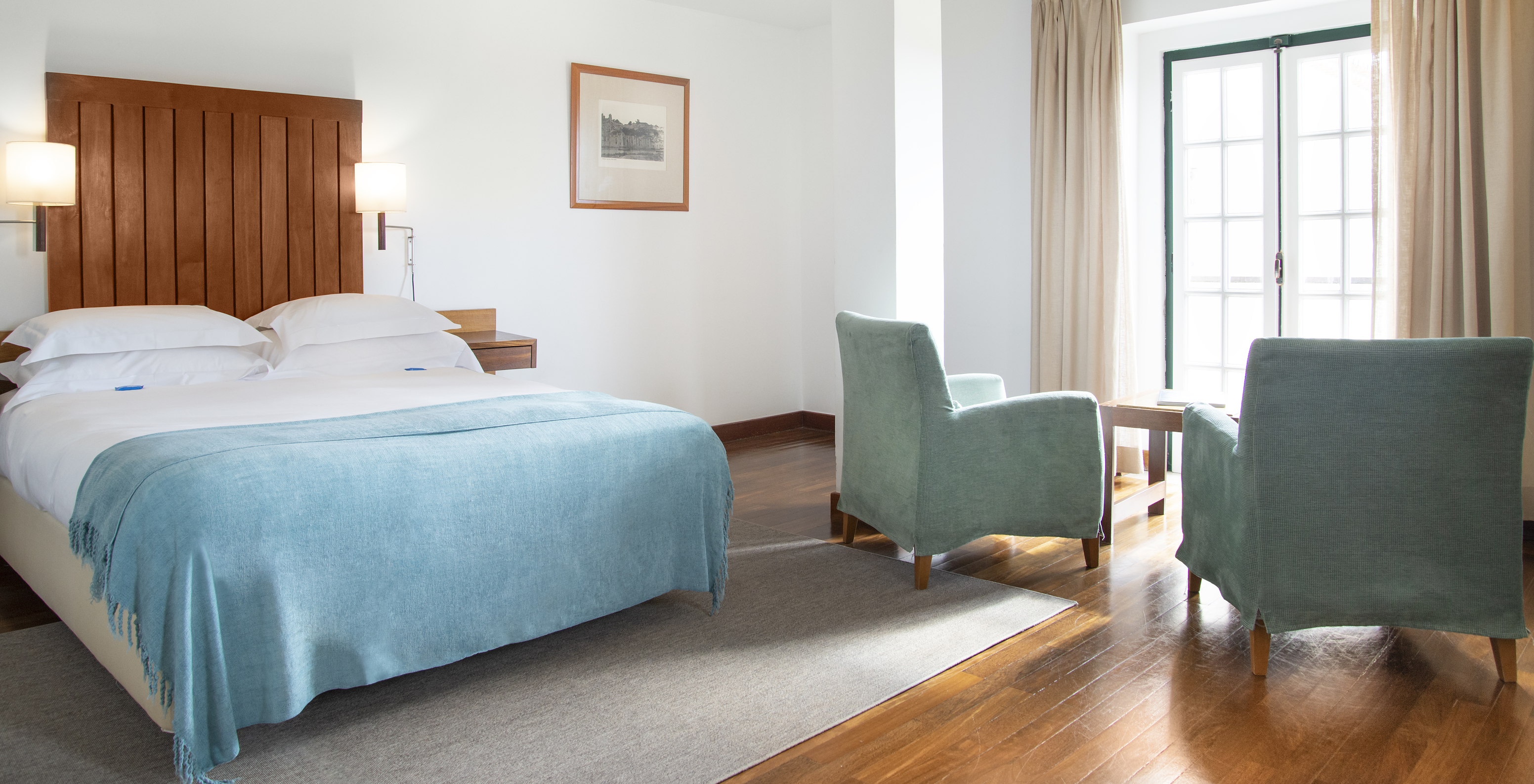 The Junior Suite of the Pousada Castelo Alcácer do Sal has a double bed with a blue blanket, two armchairs, and a window