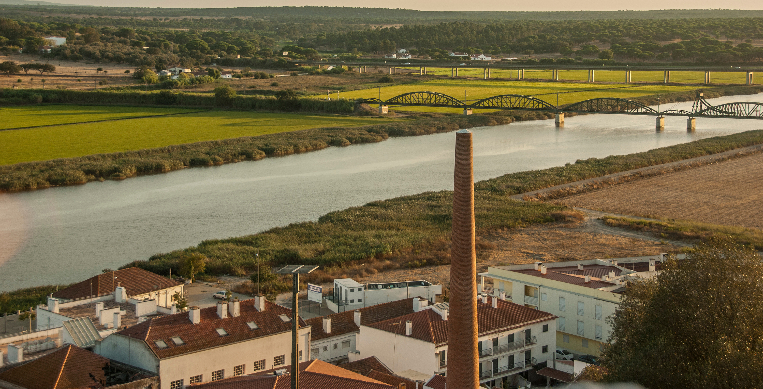 Aerial view of Alcácer do Sal, with white houses, a chimney, the Sado River, and a bridge over it