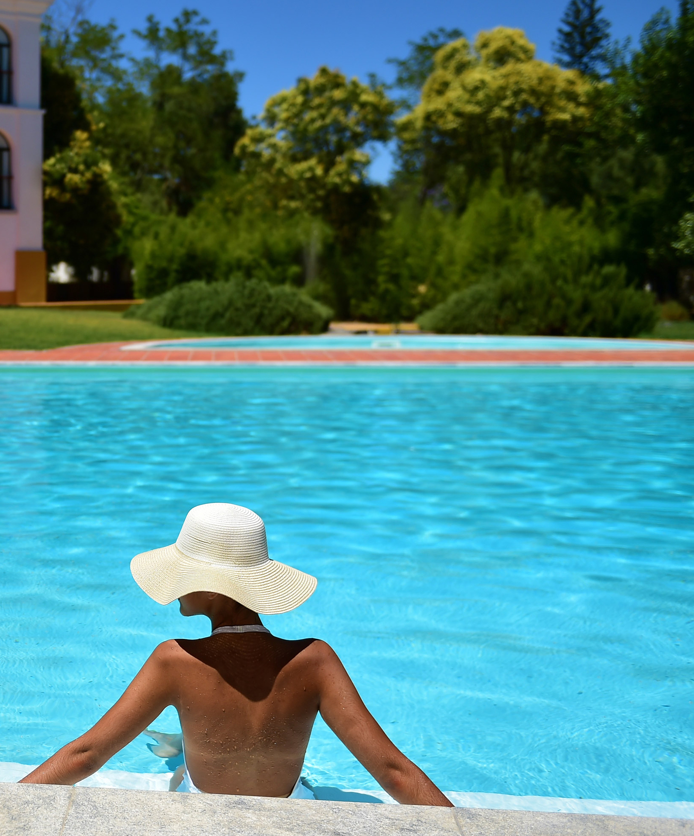 Woman with a white hat relaxing by the outdoor pool at Pousada Convento Beja, with gardens and building behind