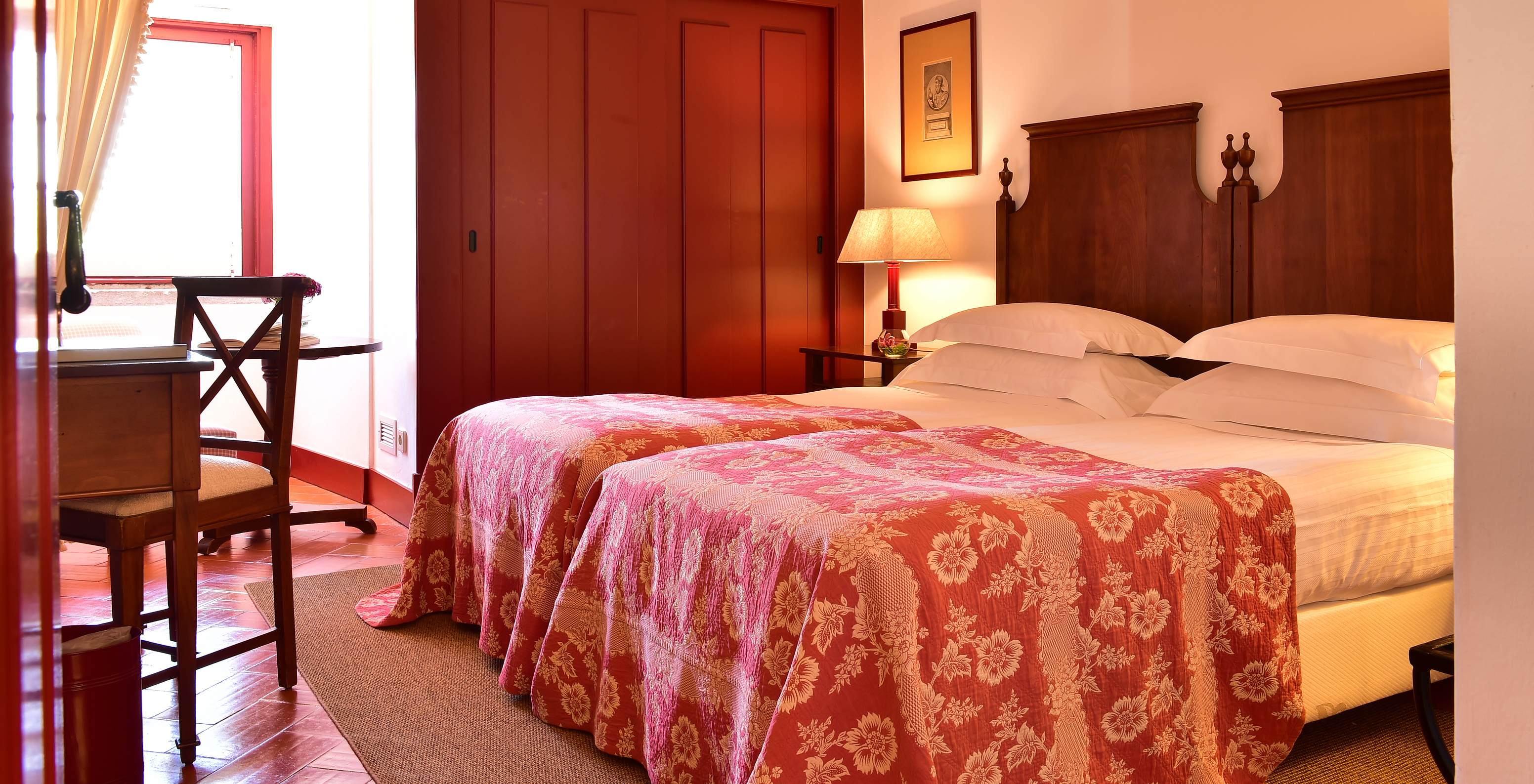 The Classic Room of The Pousada Convento Beja has two joined beds with a red bedspread, a window, and wardrobes