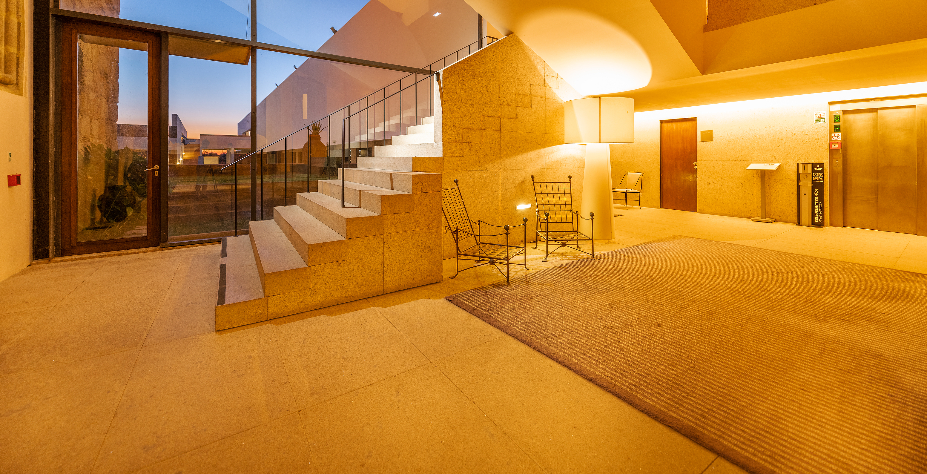 Hotel lobby with access to elevators and a large staircase leading to the rooms, and an exit door to the gardens