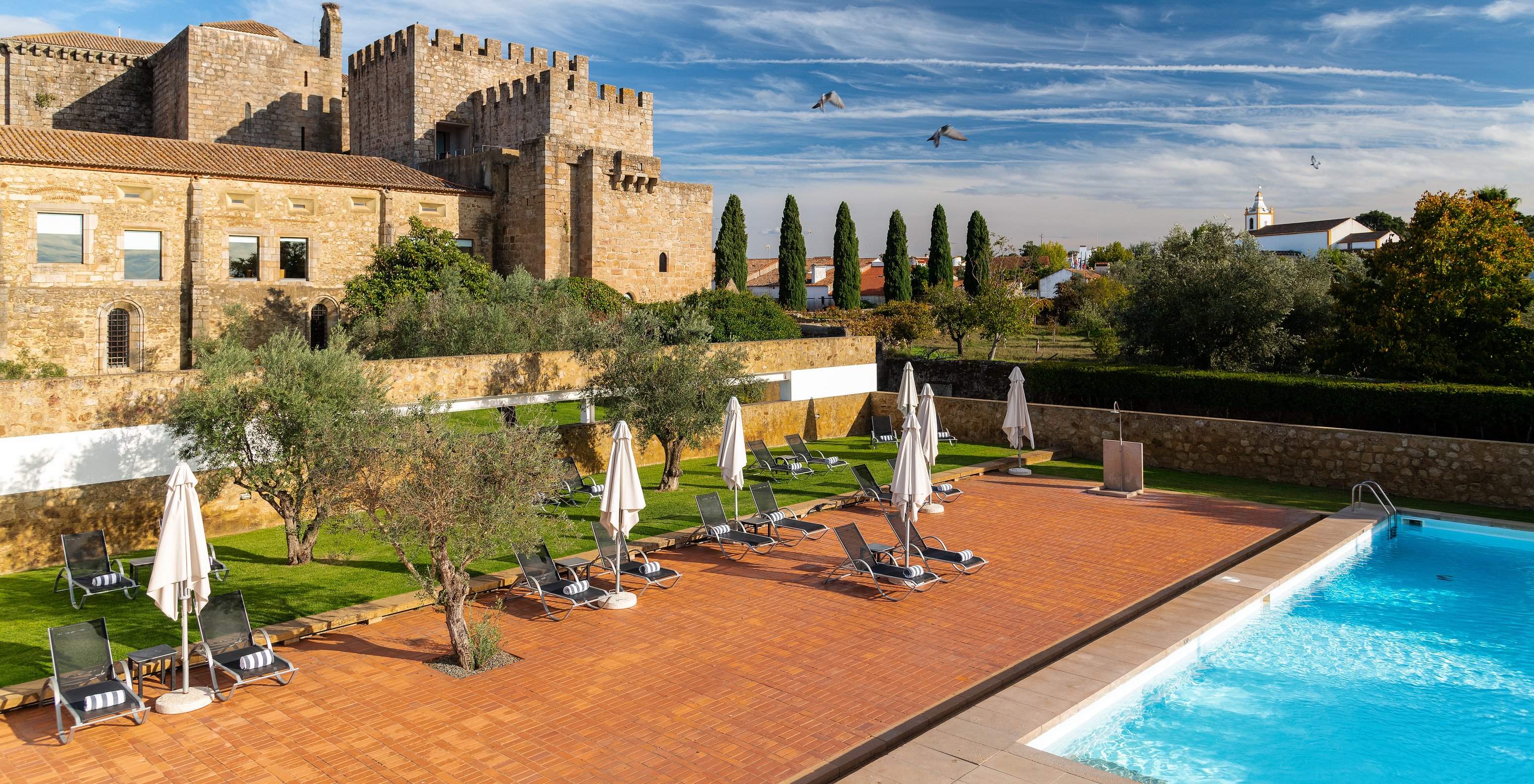 Zona de la piscina exterior del hotel con tumbonas y sombrillas, y pájaros volando en el cielo