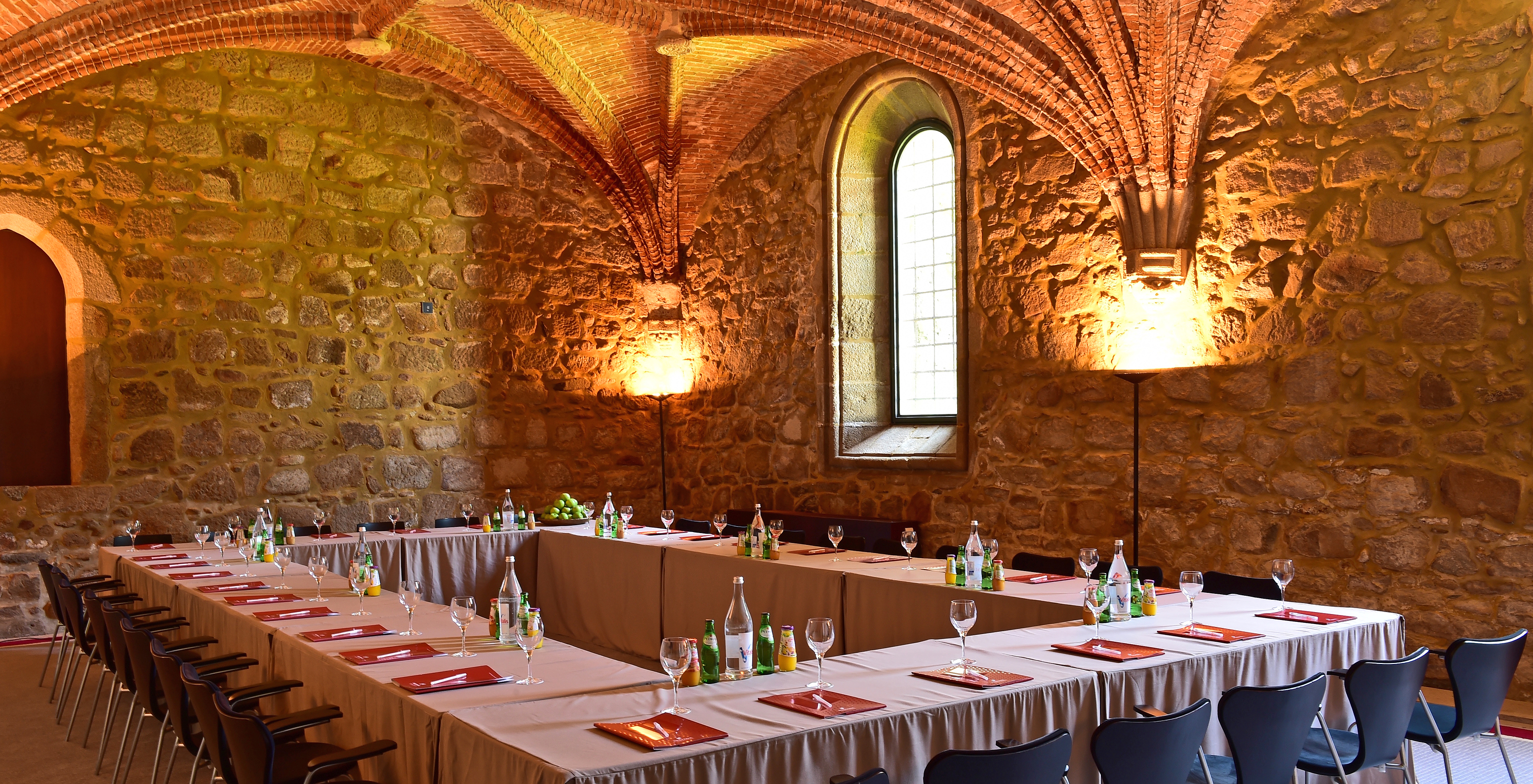 Salle de réunion avec des chaises autour d'une table, avec des cahiers et des bouteilles d'eau, sous un plafond en pierre