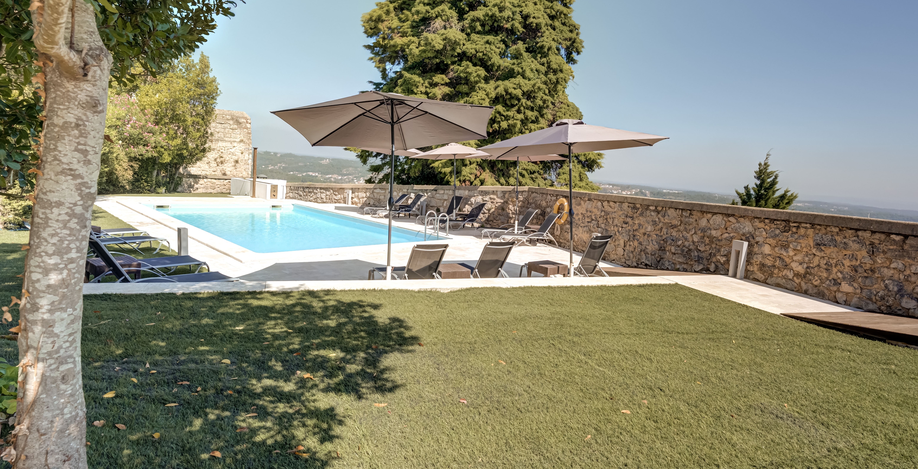 Pelouse verte près de la piscine extérieure de la Pousada Ourém, avec transats et parasols, et un arbre en arrière-plan