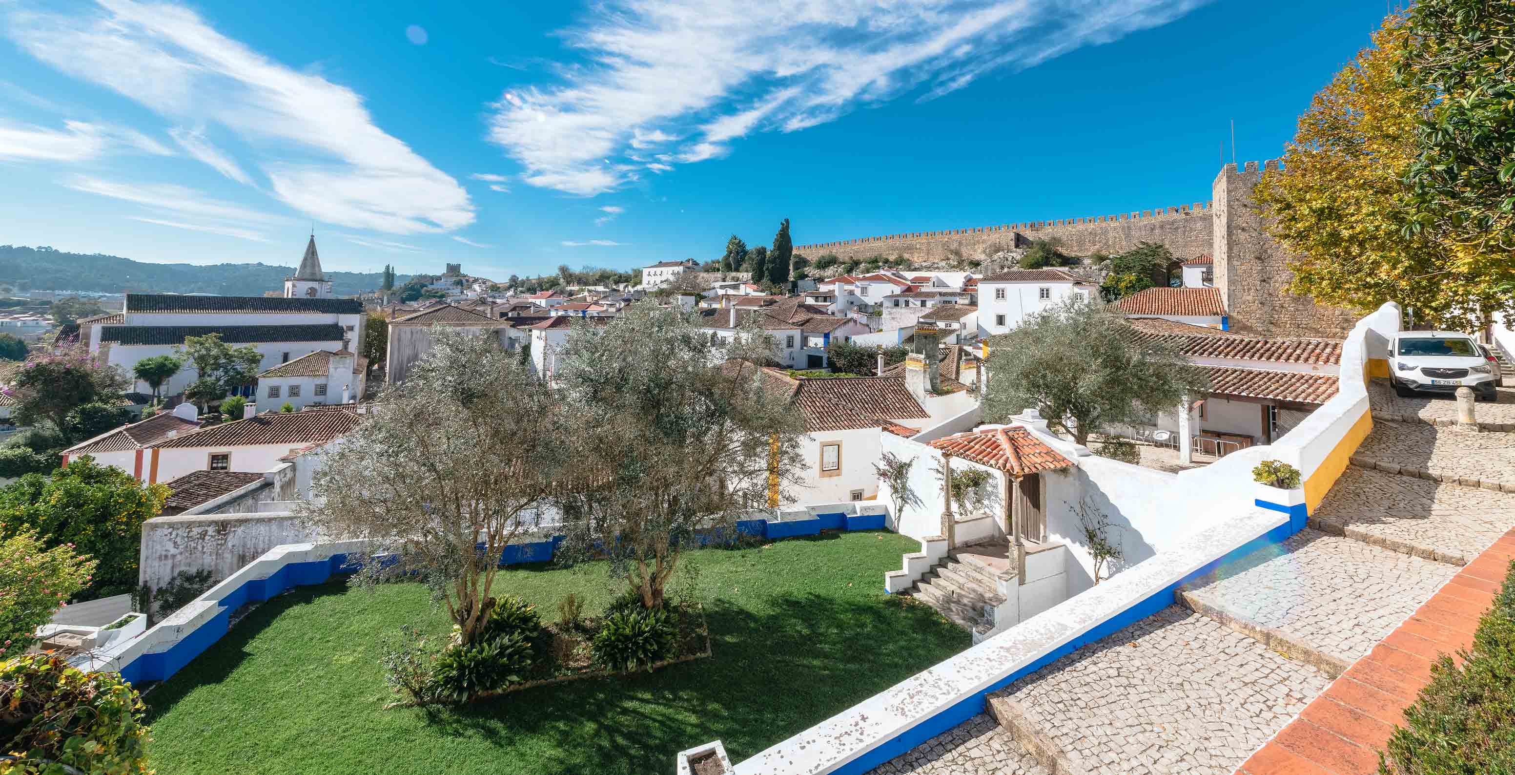 Vista do exterior da Pousada Castelo de Óbidos, com umas escadas e um pequeno jardim com a cidade ao fundo