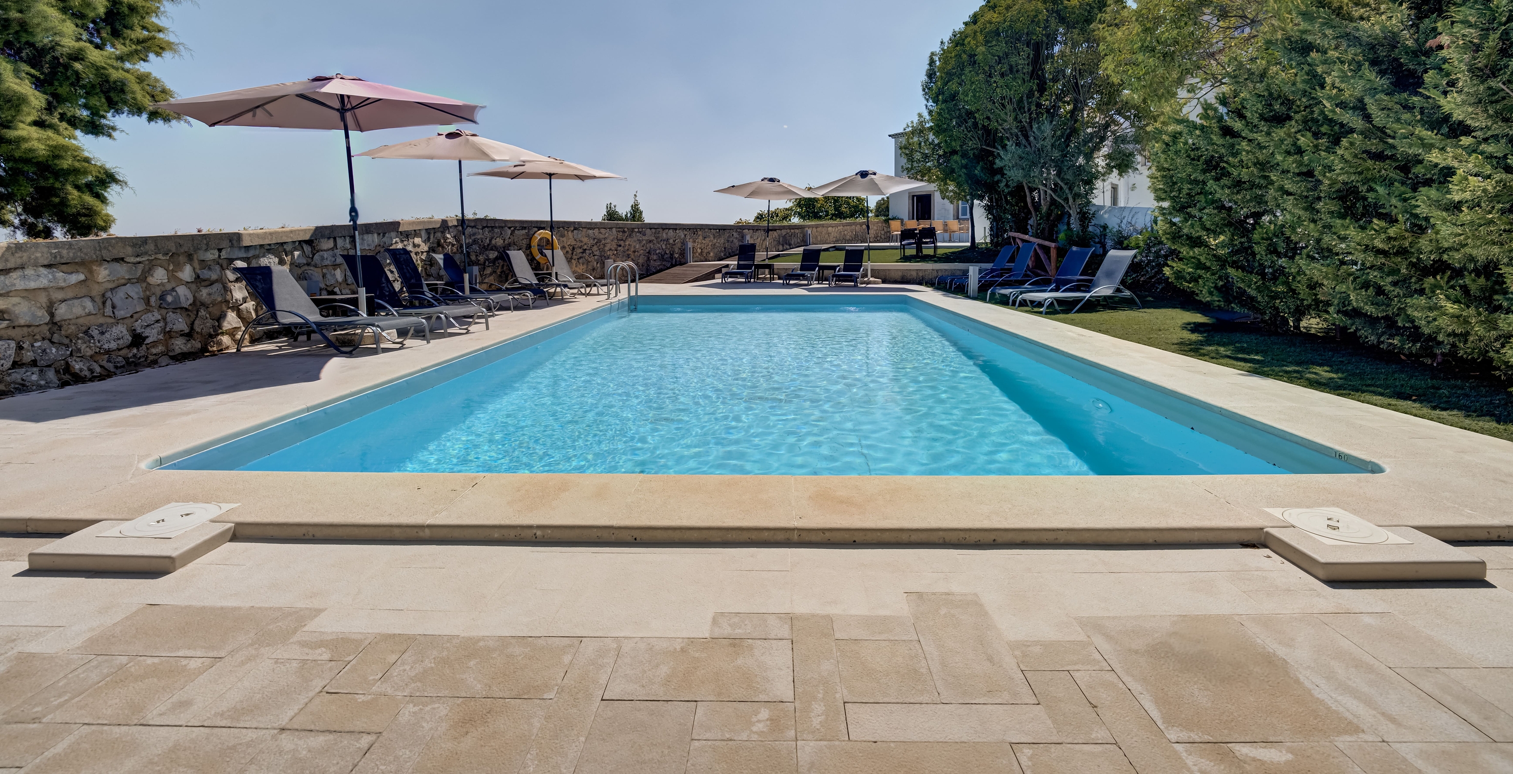 Piscina exterior, com espreguiçadeiras e chapéus de sol, com jardins verdes e várias árvores na Pousada Ourém